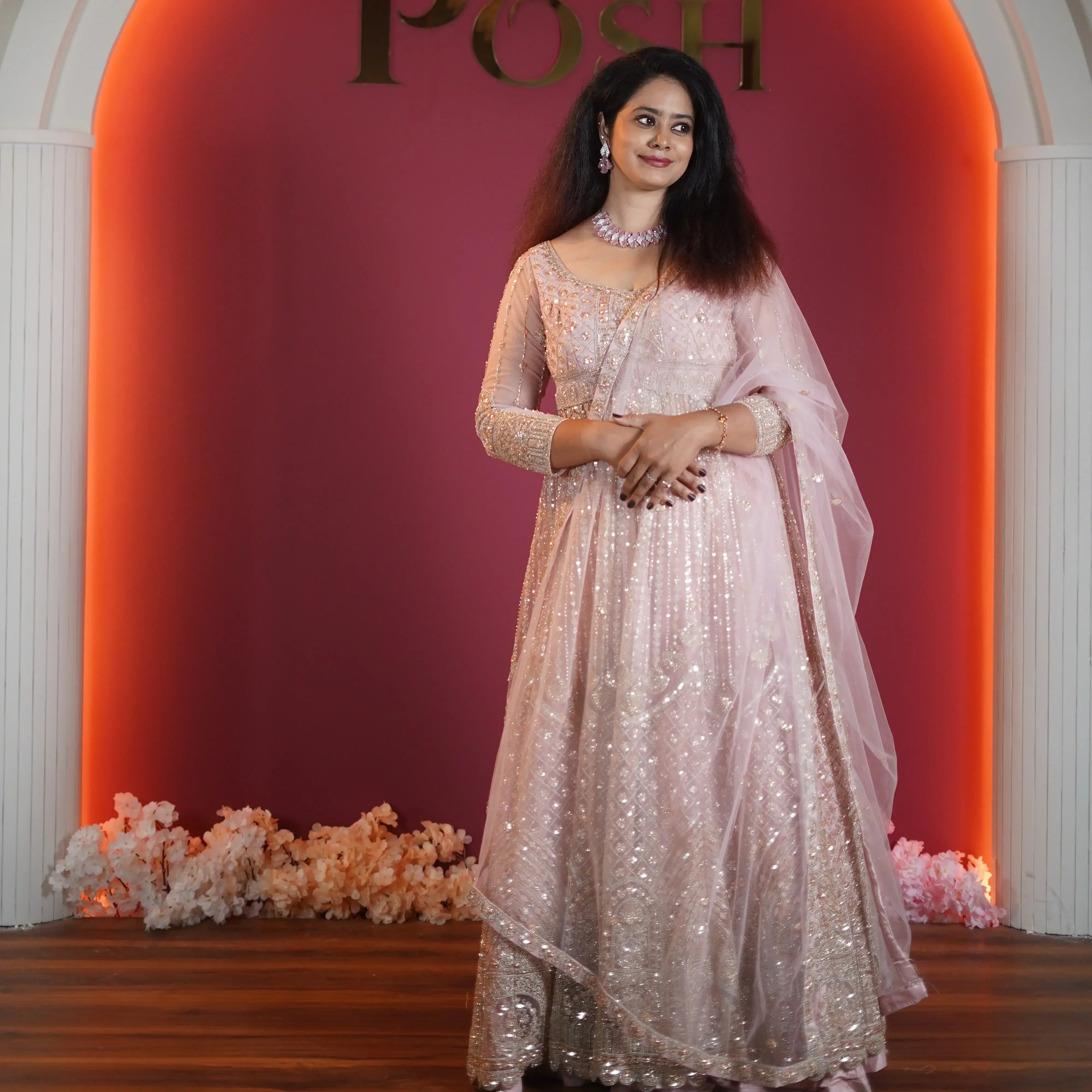 Woman wearing an elegant peach embroidered bridal gown standing under a decorative arch with warm lighting at a Posh studio setup. ✨