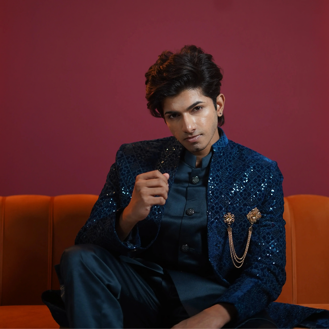 Man wearing a navy blue embroidered sherwani with gold brooch detailing, sitting on an orange sofa against a red background, posing for a fashion photoshoot.