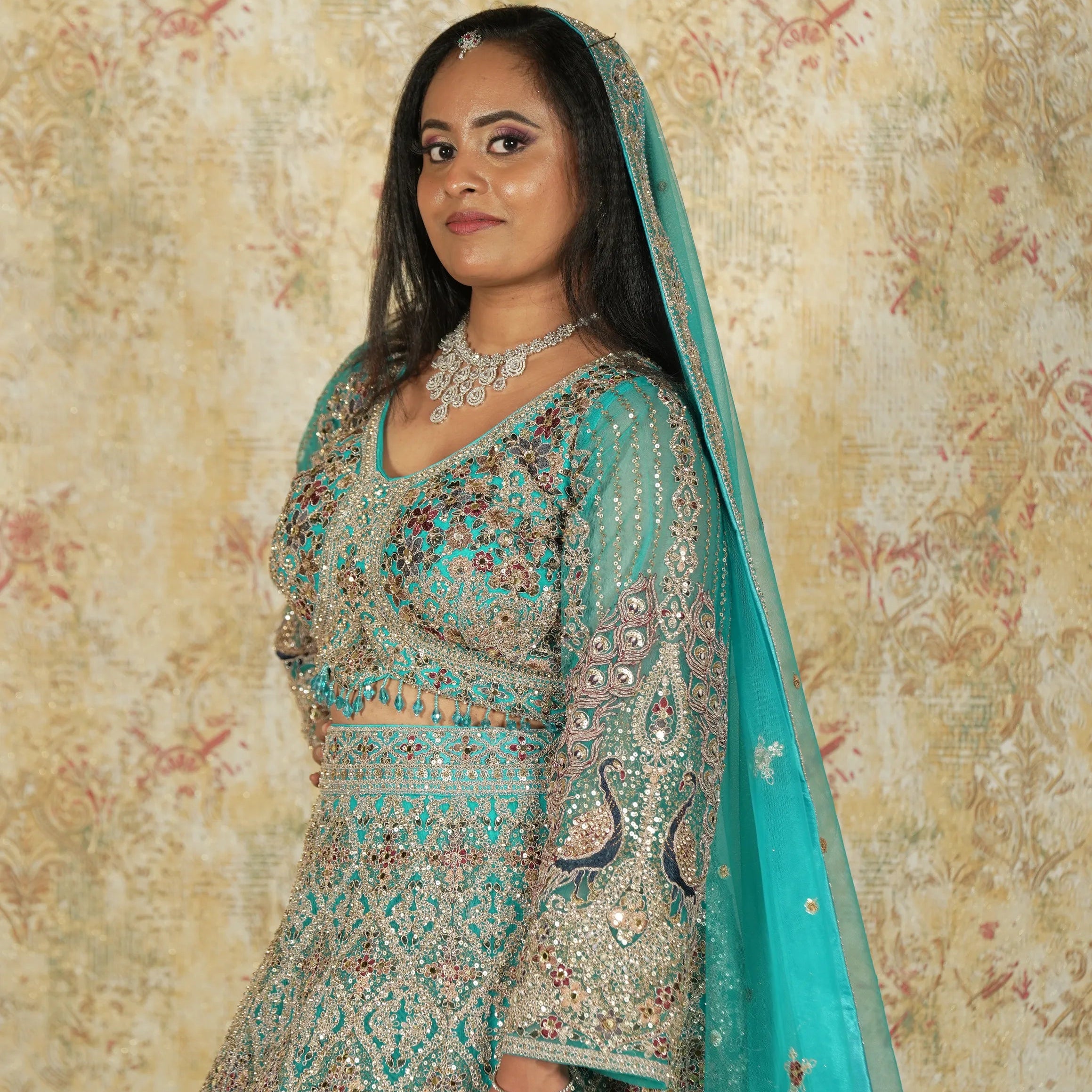 Woman in traditional embroidered outfit with teal dupatta against a patterned wall