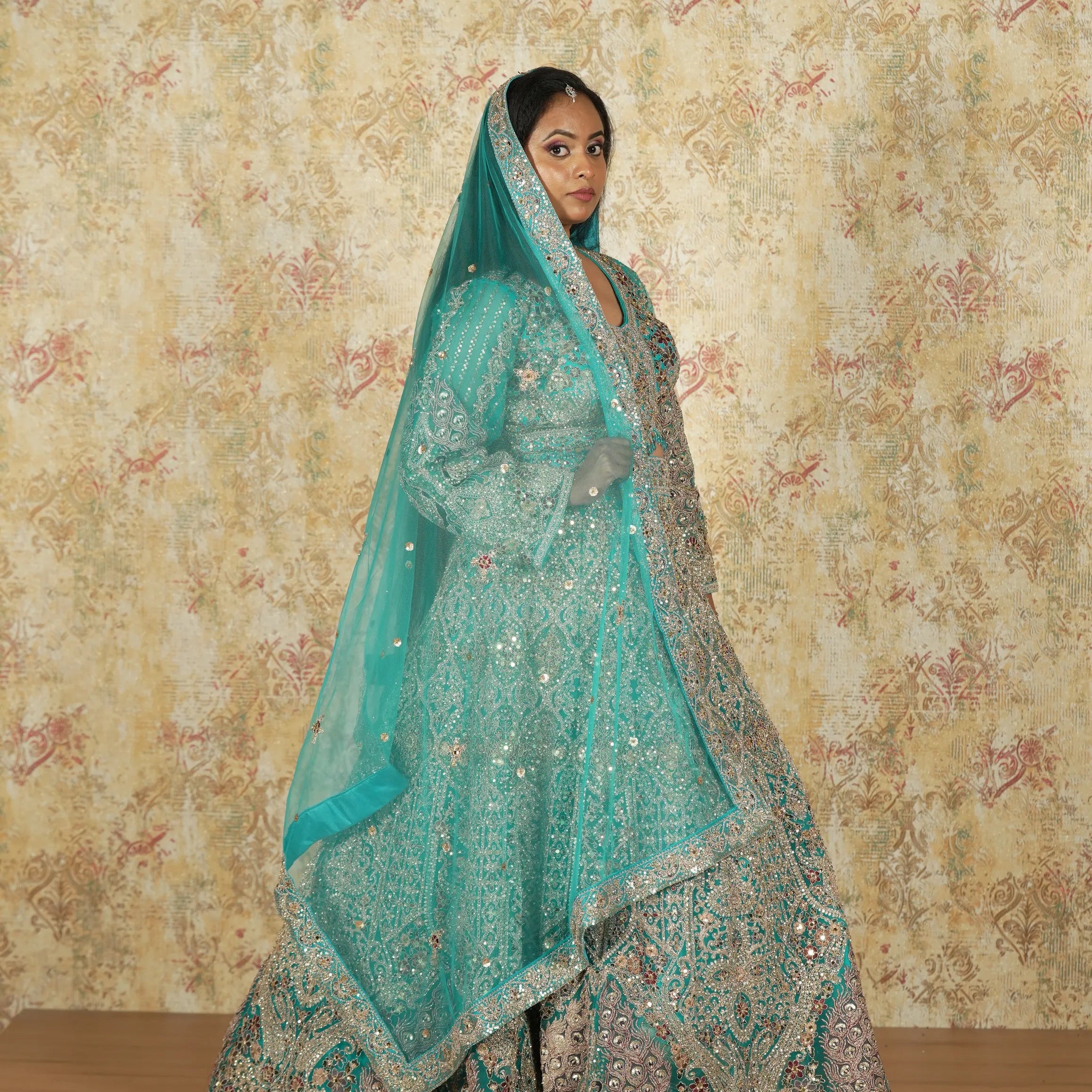Woman in a traditional turquoise embroidered outfit against a floral patterned wall.