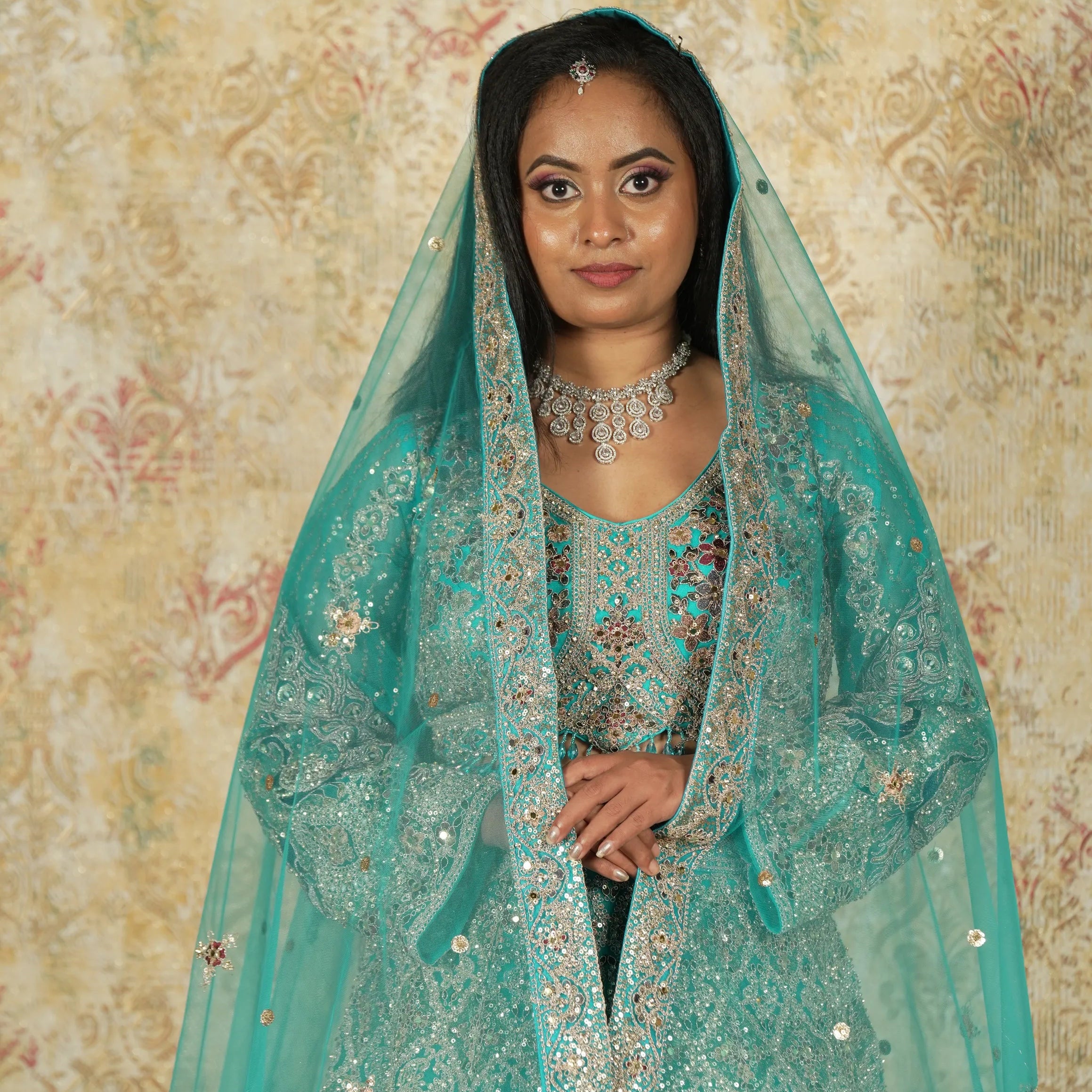 Woman in a teal embroidered outfit with a matching dupatta against a floral-patterned wall.