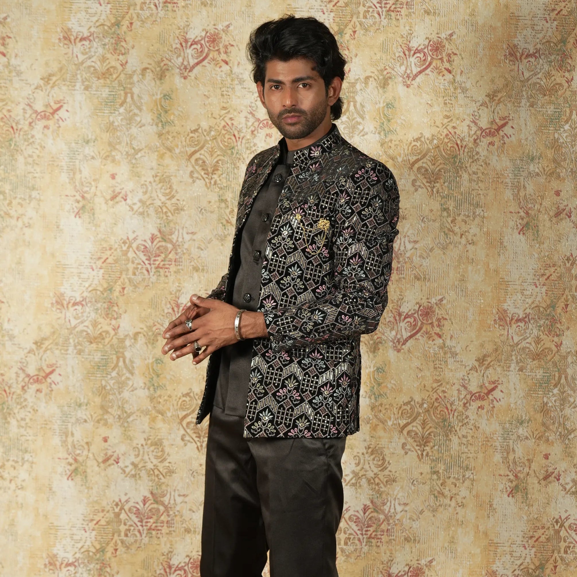 Man wearing a black embroidered jacket and pants against a patterned wall.