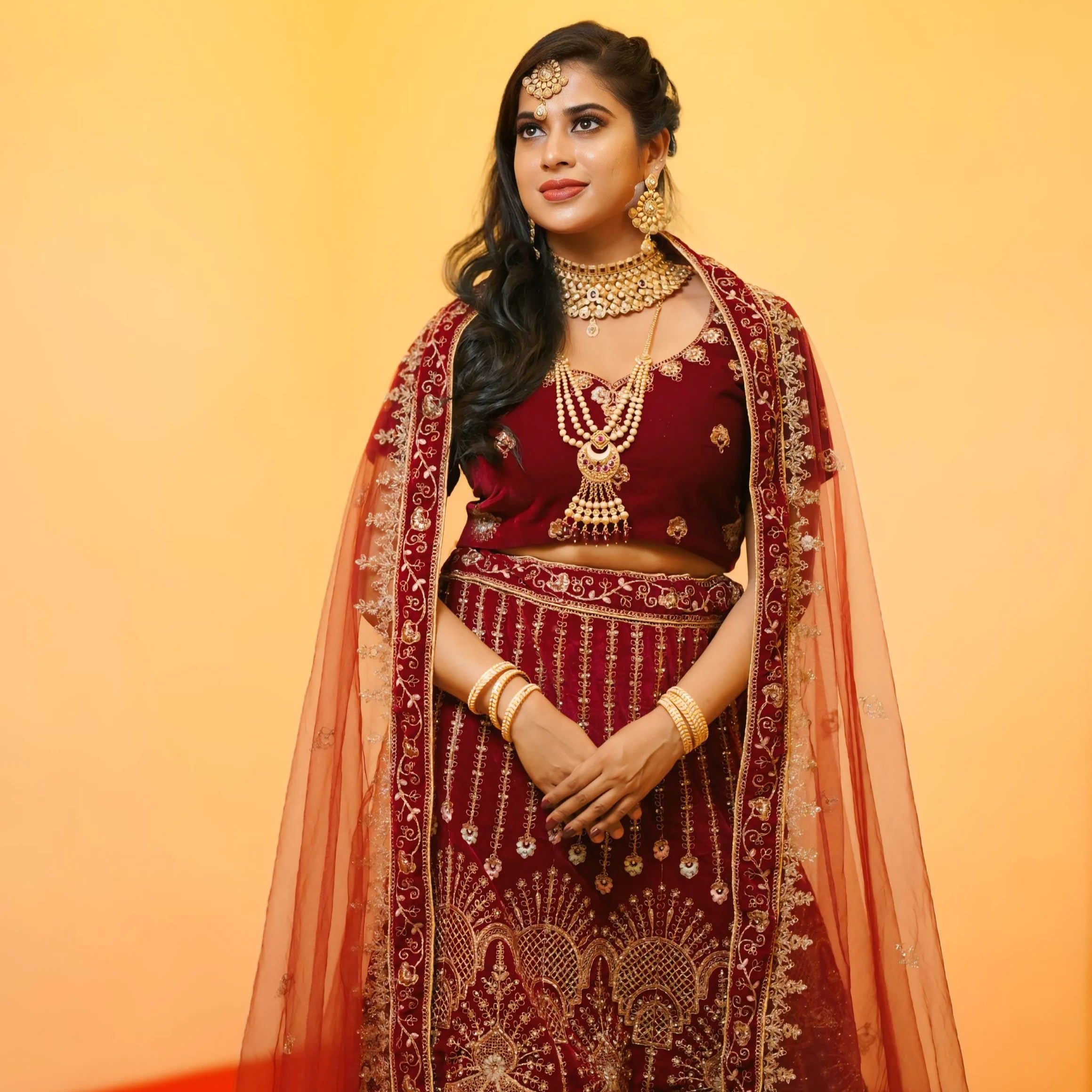 Woman in traditional maroon and gold embroidered outfit with jewelry against a warm gradient background